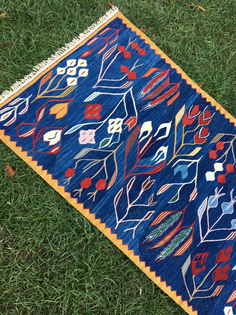 May include: A blue and yellow rug with a geometric pattern of stylized flowers and leaves. The rug has a fringe border.