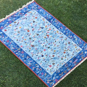 May include: A rectangular, light blue rug with a floral design. The rug features a dark blue border with a red accent and white fringe. The central design includes colorful flowers and winding vines.
