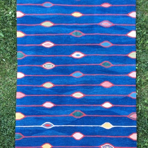 Puede incluir: Una alfombra azul con un borde de flecos blancos presenta un patr&oacute;n repetitivo de formas de ojos abstractas de colores. La alfombra tiene un dise&ntilde;o moderno y geom&eacute;trico.