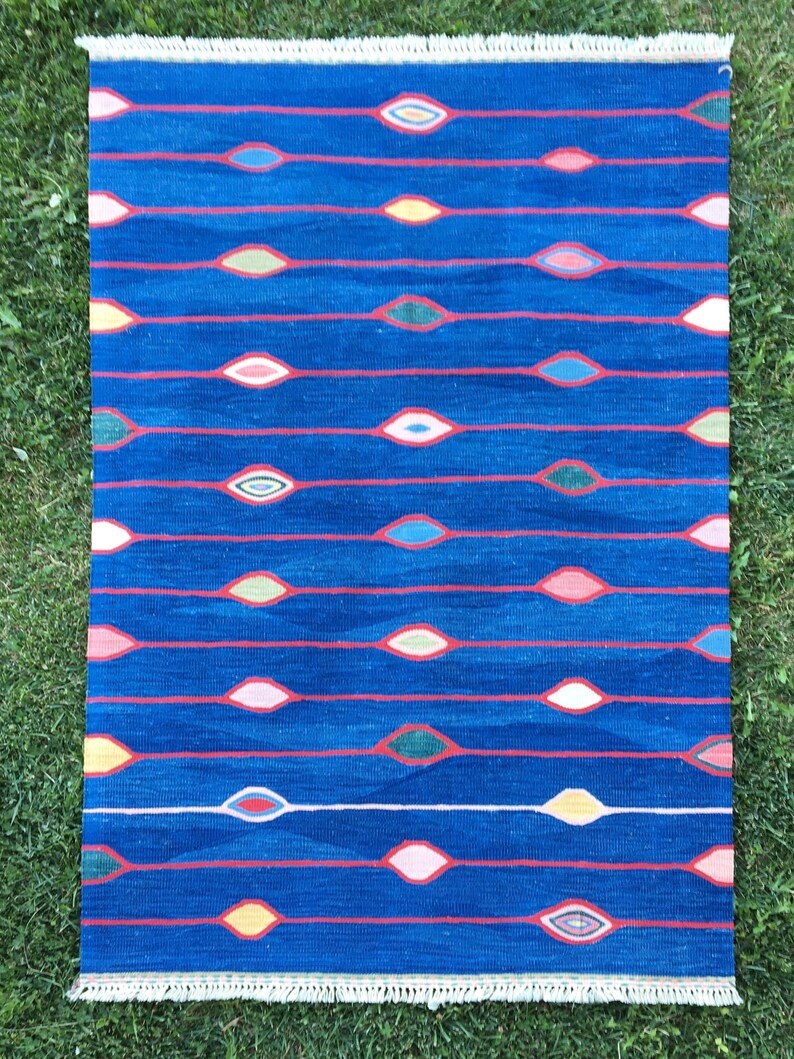 Puede incluir: Una alfombra azul con un borde de flecos blancos presenta un patr&oacute;n repetitivo de formas de ojos abstractas de colores. La alfombra tiene un dise&ntilde;o moderno y geom&eacute;trico.