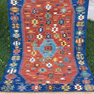 Puede incluir: Una alfombra azul y roja con un patr&oacute;n geom&eacute;trico. La alfombra tiene un borde de flecos.