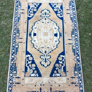May include: A vintage rug with a beige background and a blue and white geometric pattern. The rug has a fringe border.
