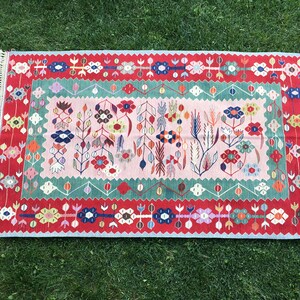 May include: Rectangular rug with a floral design. The rug features a pink center with colorful flowers and green and red borders. The edges have a white fringe.