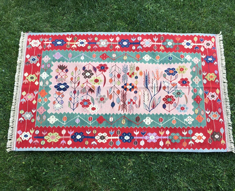 May include: Rectangular rug with a floral design. The rug features a pink center with colorful flowers and green and red borders. The edges have a white fringe.