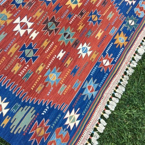 Puede incluir: Una alfombra azul y roja con un borde de flecos. La alfombra presenta un dise&ntilde;o geom&eacute;trico con estrellas, diamantes y tri&aacute;ngulos.