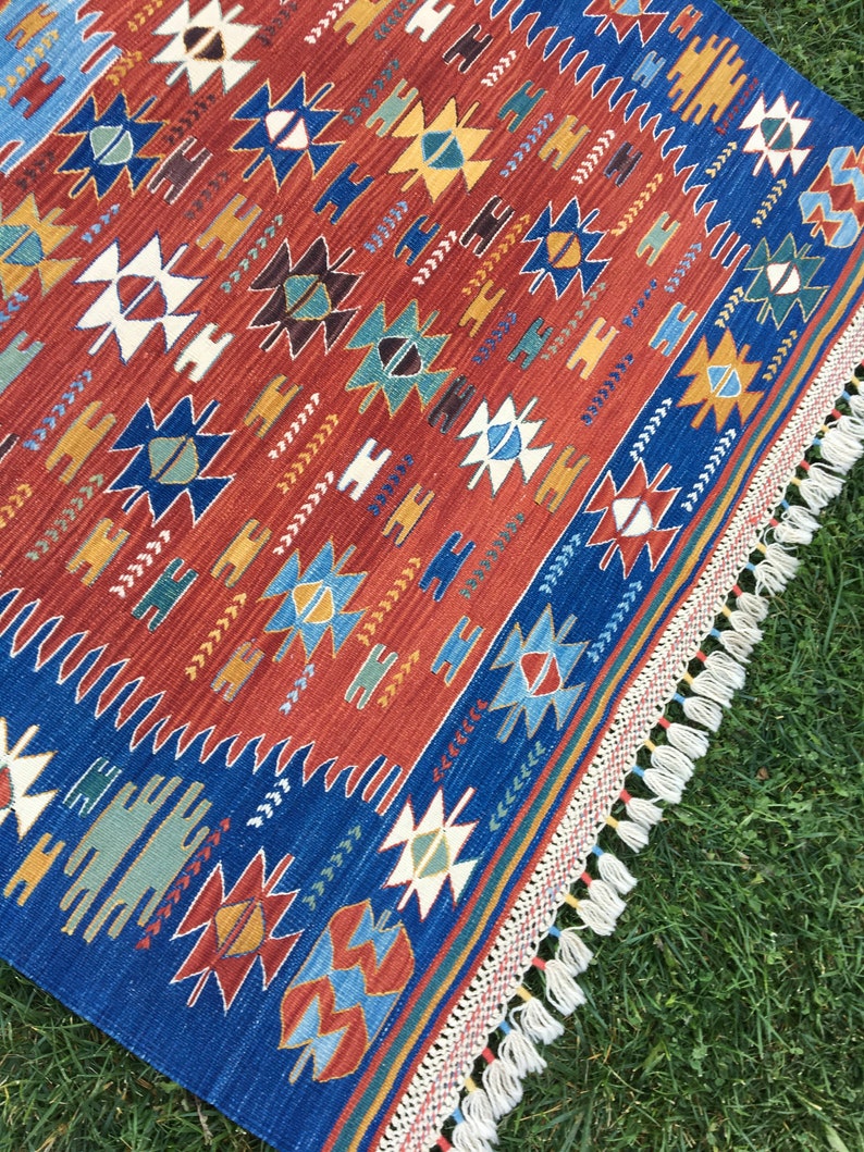 Puede incluir: Una alfombra azul y roja con un borde de flecos. La alfombra presenta un dise&ntilde;o geom&eacute;trico con estrellas, diamantes y tri&aacute;ngulos.