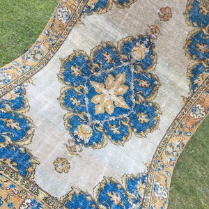 May include: An antique rug with a central medallion design in shades of blue and gold on a light beige background. The rug features intricate floral patterns and a detailed border in complementary colors. The rug is displayed outdoors on green grass.