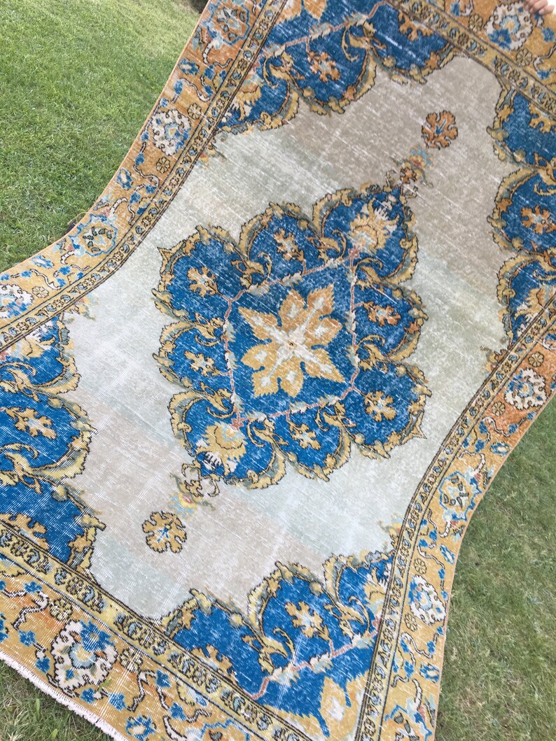 May include: An antique rug with a central medallion design in shades of blue and gold on a light beige background. The rug features intricate floral patterns and a detailed border in complementary colors. The rug is displayed outdoors on green grass.