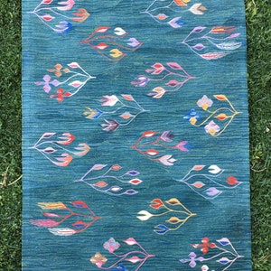 Puede incluir: Una alfombra azul verdosa con un patr&oacute;n geom&eacute;trico de flores estilizadas en tonos de rosa, morado, azul, rojo, amarillo y blanco. La alfombra tiene un borde de flecos.