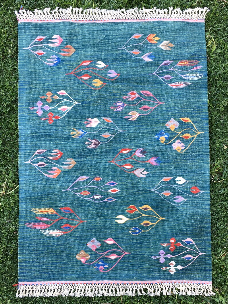 Puede incluir: Una alfombra azul verdosa con un patr&oacute;n geom&eacute;trico de flores estilizadas en tonos de rosa, morado, azul, rojo, amarillo y blanco. La alfombra tiene un borde de flecos.