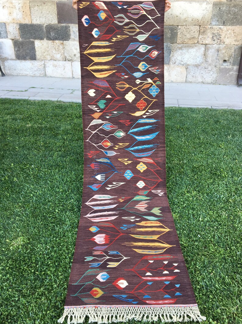 Puede incluir: Una alfombra larga y estrecha con fondo marr&oacute;n y un patr&oacute;n geom&eacute;trico colorido. El patr&oacute;n presenta hojas y flores estilizadas en tonos de azul, rojo, amarillo y verde. La alfombra tiene flecos en ambos extremos.