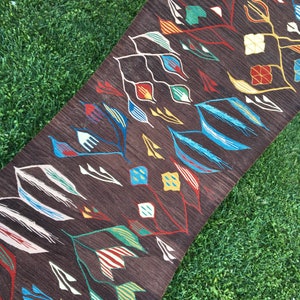 Puede incluir: Una alfombra marr&oacute;n con un patr&oacute;n geom&eacute;trico en tonos de azul, amarillo, rojo, verde y blanco. El patr&oacute;n presenta hojas estilizadas y diamantes.