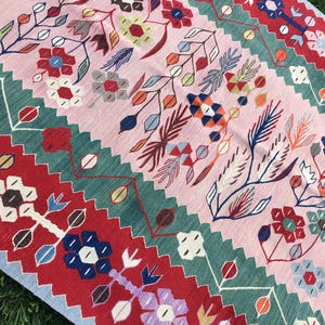 May include: A colorful, floral-patterned rug with a pink background and vibrant accents of red, green, blue, and white. The rug features a repeating design of stylized flowers and leaves, creating a lively and decorative textile.