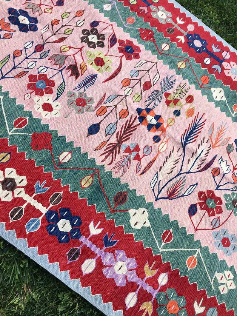 May include: A colorful, floral-patterned rug with a pink background and vibrant accents of red, green, blue, and white. The rug features a repeating design of stylized flowers and leaves, creating a lively and decorative textile.