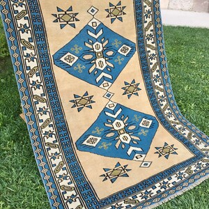 May include: A beige rug with a blue and green geometric pattern. The rug has a fringe border.