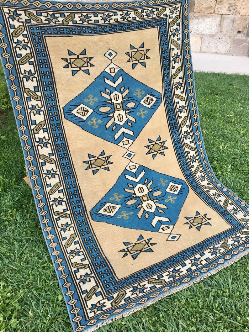 May include: A beige rug with a blue and green geometric pattern. The rug has a fringe border.