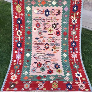 May include: A rectangular, hand-woven rug with a vibrant floral design. The rug features a red border, a central pink field, and accents of green, blue, and orange. The design includes stylized flowers and leaf patterns, with a fringed edge.