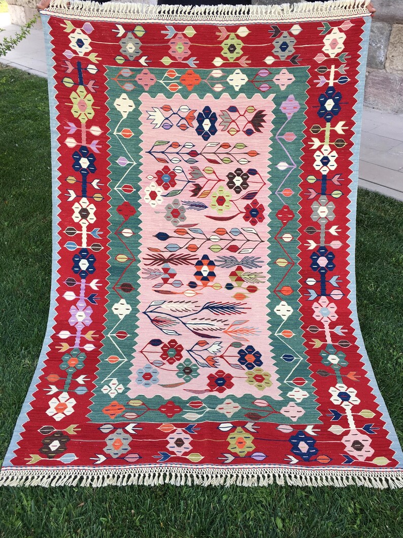 May include: A rectangular, hand-woven rug with a vibrant floral design. The rug features a red border, a central pink field, and accents of green, blue, and orange. The design includes stylized flowers and leaf patterns, with a fringed edge.