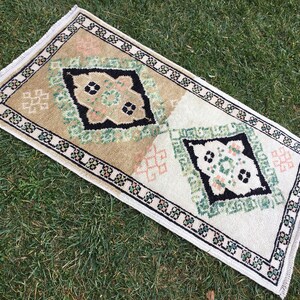 May include: A small, rectangular rug with a white background and a black, green, and pink geometric pattern. The rug has a fringed border.
