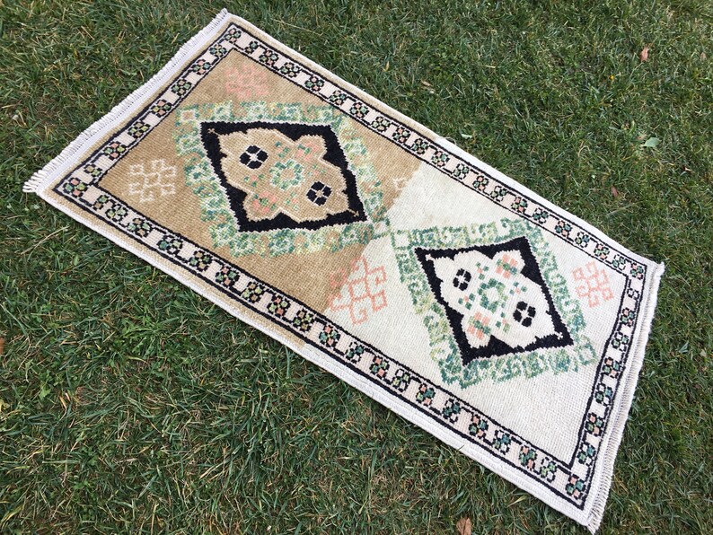 May include: A small, rectangular rug with a white background and a black, green, and pink geometric pattern. The rug has a fringed border.