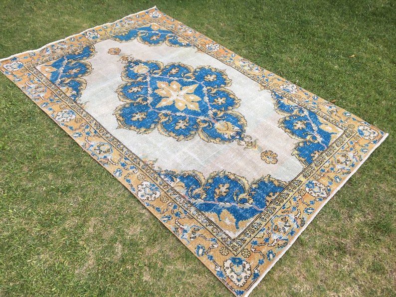 May include: Rectangular area rug with a central medallion design in blue and gold against a cream background. The rug has an ornate border with floral patterns. The rug is placed on green grass.