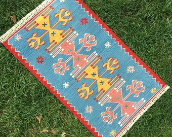 May include: A blue, red, and yellow patterned rug with a fringe border. The rug has a geometric design with a repeating pattern of stylized flowers.