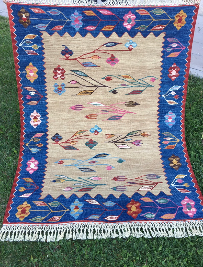 May include: A beige rug with a blue border and a pattern of stylized leaves and flowers in shades of pink, blue, red, and green. The rug has a fringe border.