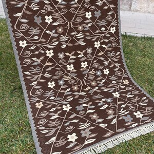 Puede incluir: Una alfombra marr&oacute;n y blanca con estampado y borde de flecos. La alfombra presenta un dise&ntilde;o floral con hojas y flores.