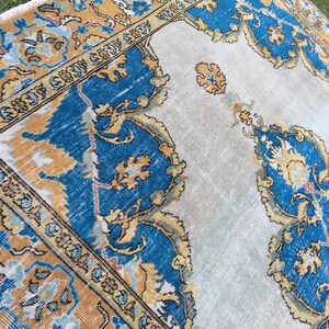 May include: A vintage-style rug with a central light beige field and ornate borders. The borders feature a combination of blue, gold, and light blue floral patterns. The rug's design includes intricate details and a distressed appearance, suggesting age and use.