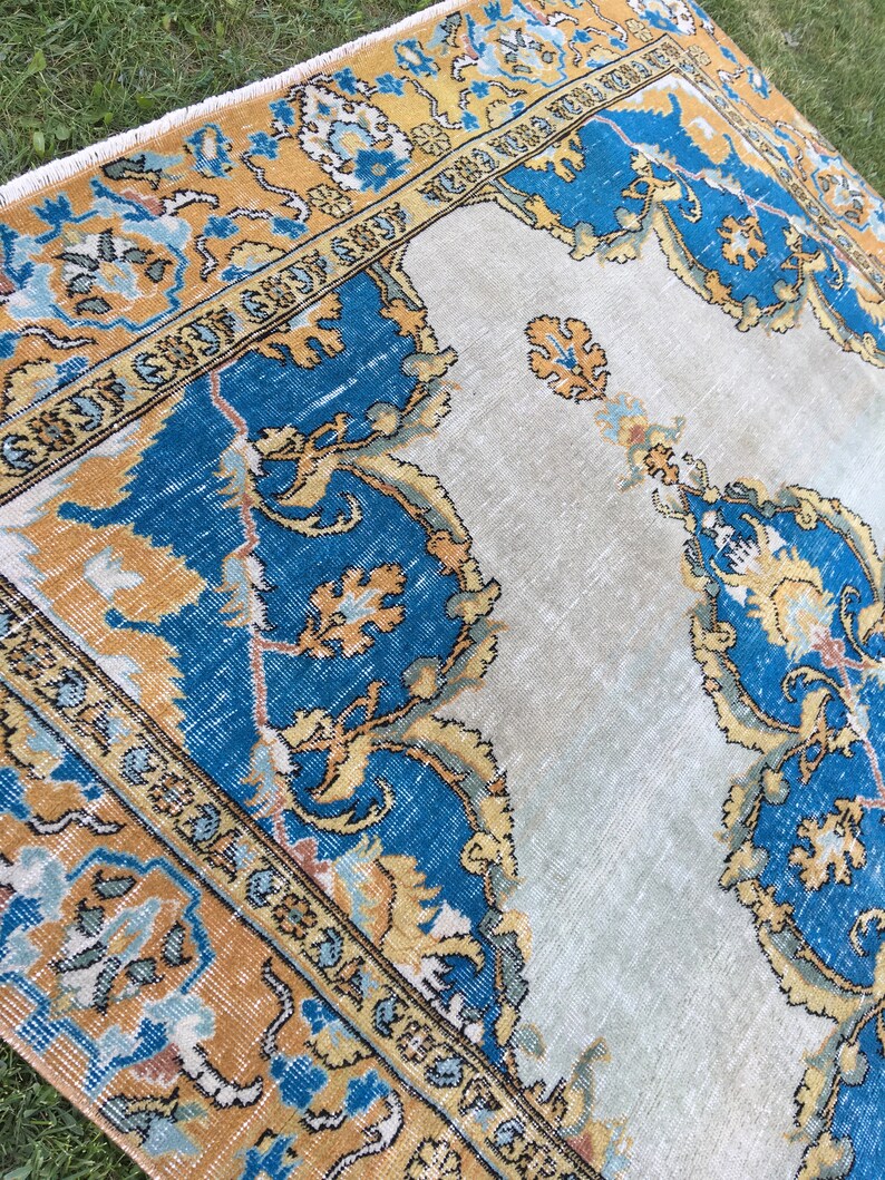 May include: A vintage-style rug with a central light beige field and ornate borders. The borders feature a combination of blue, gold, and light blue floral patterns. The rug's design includes intricate details and a distressed appearance, suggesting age and use.