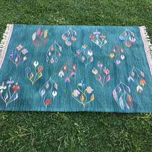 Puede incluir: Una alfombra azul turquesa con un patr&oacute;n floral en tonos de rosa, amarillo, naranja y blanco. La alfombra tiene un borde de flecos.