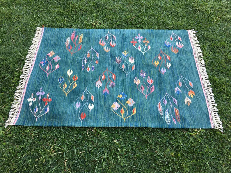 Puede incluir: Una alfombra azul turquesa con un patr&oacute;n floral en tonos de rosa, amarillo, naranja y blanco. La alfombra tiene un borde de flecos.