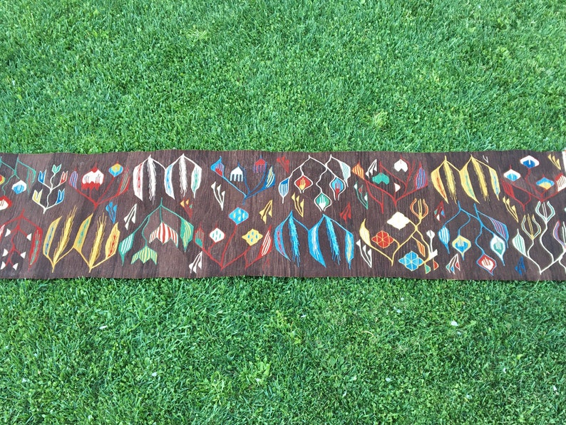 Puede incluir: Una alfombra larga y estrecha con fondo marr&oacute;n y un patr&oacute;n geom&eacute;trico colorido. El patr&oacute;n presenta flores y hojas estilizadas en tonos de rojo, azul, amarillo, verde y blanco.