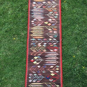 Puede incluir: Una alfombra larga y estrecha con fondo marr&oacute;n y un estampado floral colorido. La alfombra tiene un borde rojo y flecos blancos en los extremos.