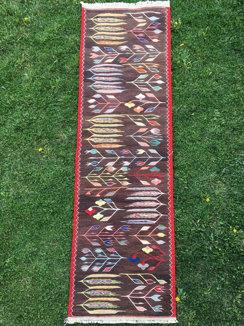 Puede incluir: Una alfombra larga y estrecha con fondo marr&oacute;n y un estampado floral colorido. La alfombra tiene un borde rojo y flecos blancos en los extremos.