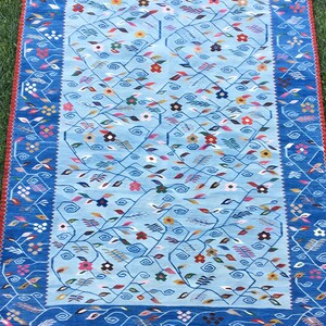 May include: A rectangular blue rug with a floral pattern. The rug has a light blue center with a darker blue border. The border features a repeating floral and leaf design in various colors, including red, pink, and yellow. The rug is placed on green grass.