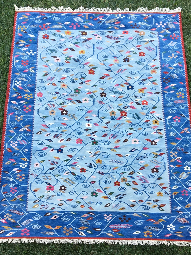 May include: A rectangular blue rug with a floral pattern. The rug has a light blue center with a darker blue border. The border features a repeating floral and leaf design in various colors, including red, pink, and yellow. The rug is placed on green grass.