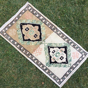 May include: A small, rectangular rug with a white background and a geometric pattern in shades of green, black, and pink. The rug has a fringed border.