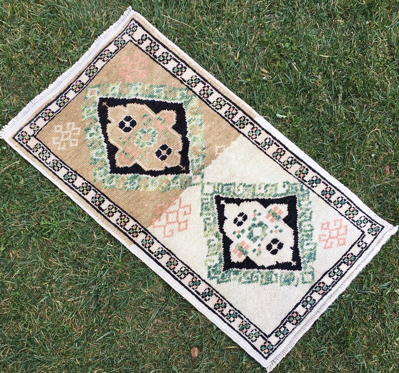 May include: A small, rectangular rug with a white background and a geometric pattern in shades of green, black, and pink. The rug has a fringed border.