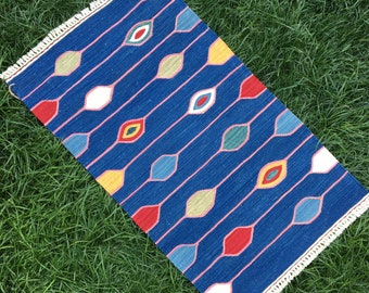 May include: A blue rug with a white fringe. The rug has a repeating pattern of colorful, abstract shapes.