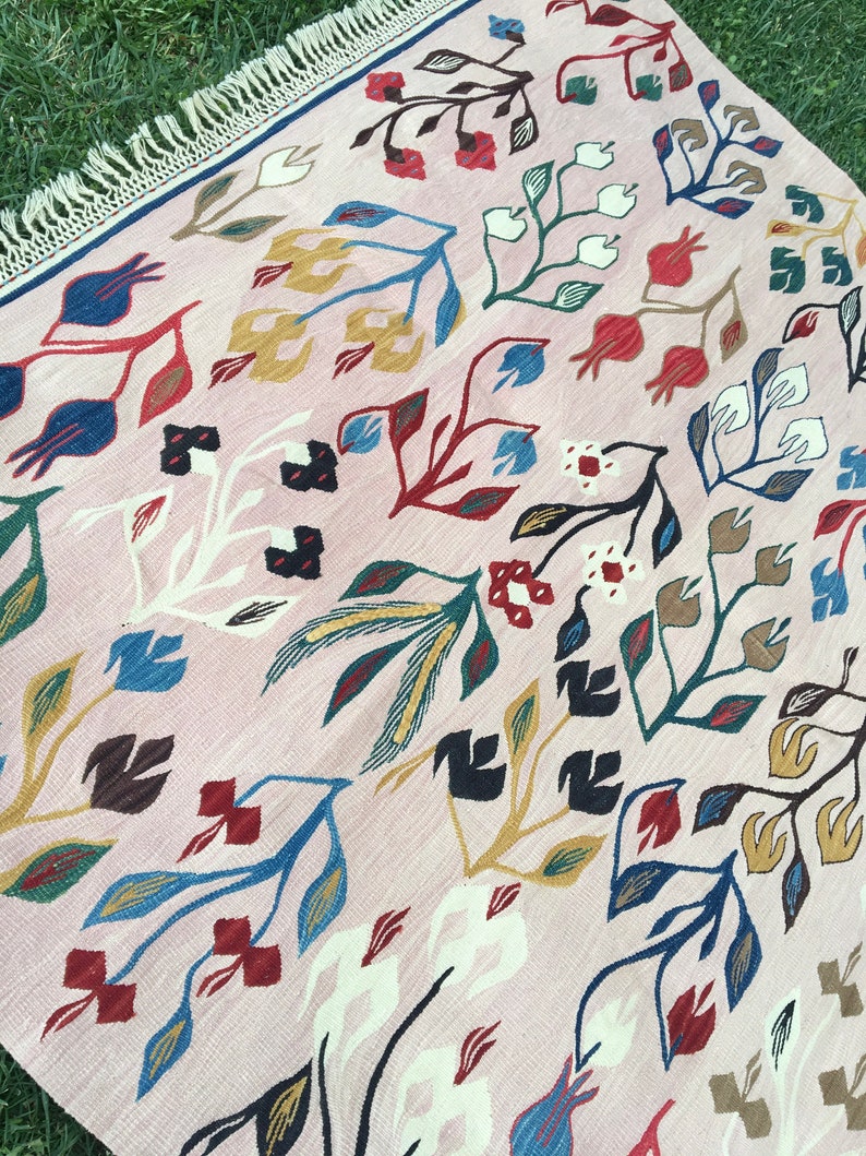 Puede incluir: Una alfombra rosa con un patr&oacute;n floral en tonos de azul, rojo, amarillo y marr&oacute;n. La alfombra tiene un borde de flecos.