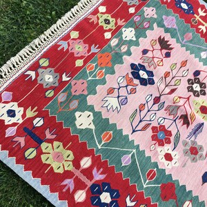 May include: A colorful, rectangular rug with a floral pattern. The rug features a red border with repeating floral designs, a central pink field, and green and blue accents. The rug has a fringed edge and is placed on a bed of green grass.