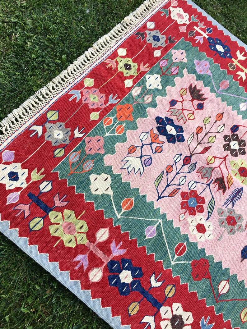 May include: A colorful, rectangular rug with a floral pattern. The rug features a red border with repeating floral designs, a central pink field, and green and blue accents. The rug has a fringed edge and is placed on a bed of green grass.