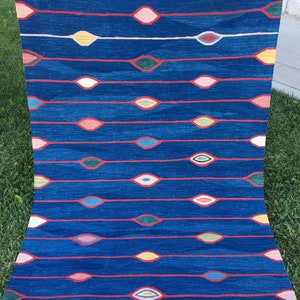 Puede incluir: Una alfombra azul con un patr&oacute;n repetitivo de dise&ntilde;os coloridos en forma de ojo. La alfombra tiene un borde de flecos.