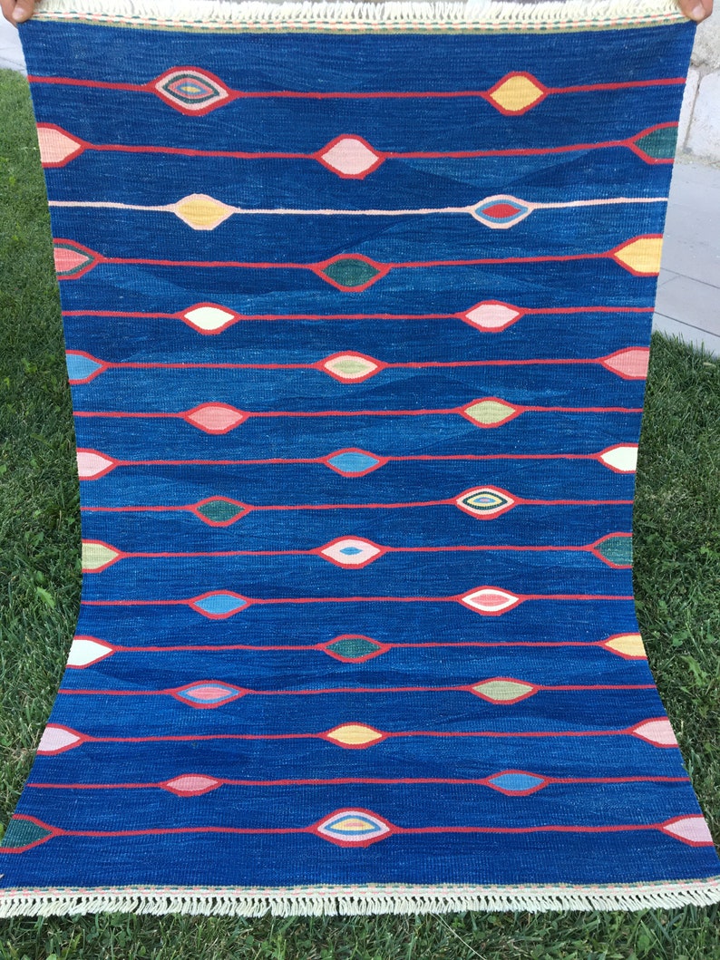 Puede incluir: Una alfombra azul con un patr&oacute;n repetitivo de dise&ntilde;os coloridos en forma de ojo. La alfombra tiene un borde de flecos.