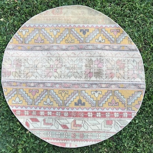Könnte beinhalten: Ein runder, cremefarbener Teppich mit einem geometrischen Muster in Rosa-, Gelb- und Grautönen. Der Teppich hat einen Fransenrand.