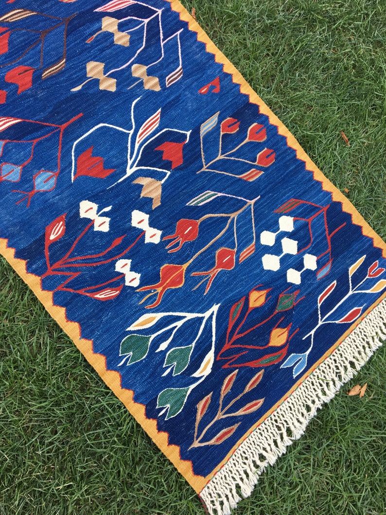 May include: A blue and yellow patterned rug with a fringe border. The rug features a stylized floral design with red, white, and green accents.