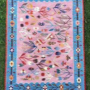 Op de afbeelding: Een roze en blauw tapijt met een bloemenpatroon. Het tapijt heeft een franje rand en is geweven met ingewikkelde details.