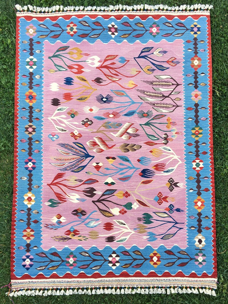 Op de afbeelding: Een roze en blauw tapijt met een bloemenpatroon. Het tapijt heeft een franje rand en is geweven met ingewikkelde details.