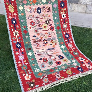 May include: A rectangular, hand-woven rug with a floral design. The rug features a red border with colorful floral motifs, a central pink field, and green and blue accents. The rug has a fringed edge and is displayed on green grass.